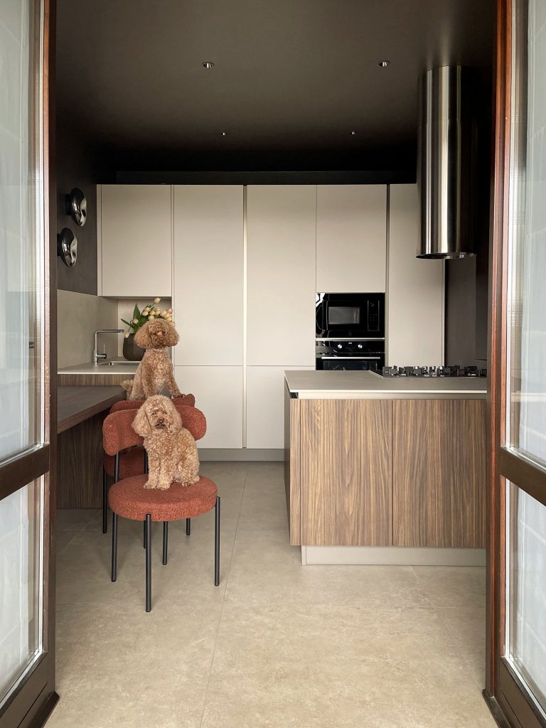 Interno 16: cucina con ante chiare e isola in legno noce, con due barboncini sulle sedie in bouclé terracotta