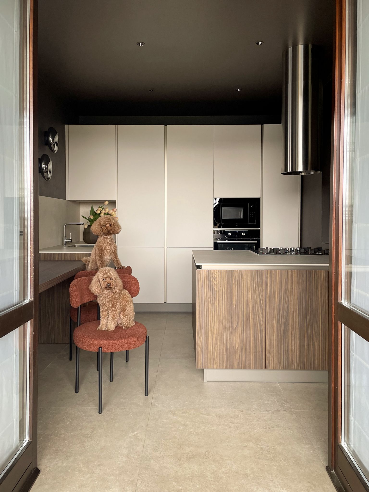 Interno 16: cucina con ante chiare e isola in legno noce, con due barboncini sulle sedie in bouclé terracotta